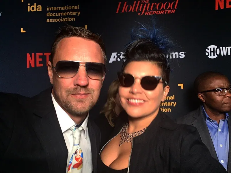 Una selfie de dos personas frente al fondo de una premiación. Detrás hay una tercera persona.