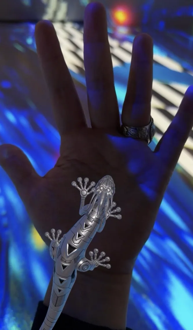 Una gecko robótica plateada sobre una mano abierta, en luz azul. Las Awichas en realidad aumentada. Personaje animado en 3D de Brian Condori Marza.