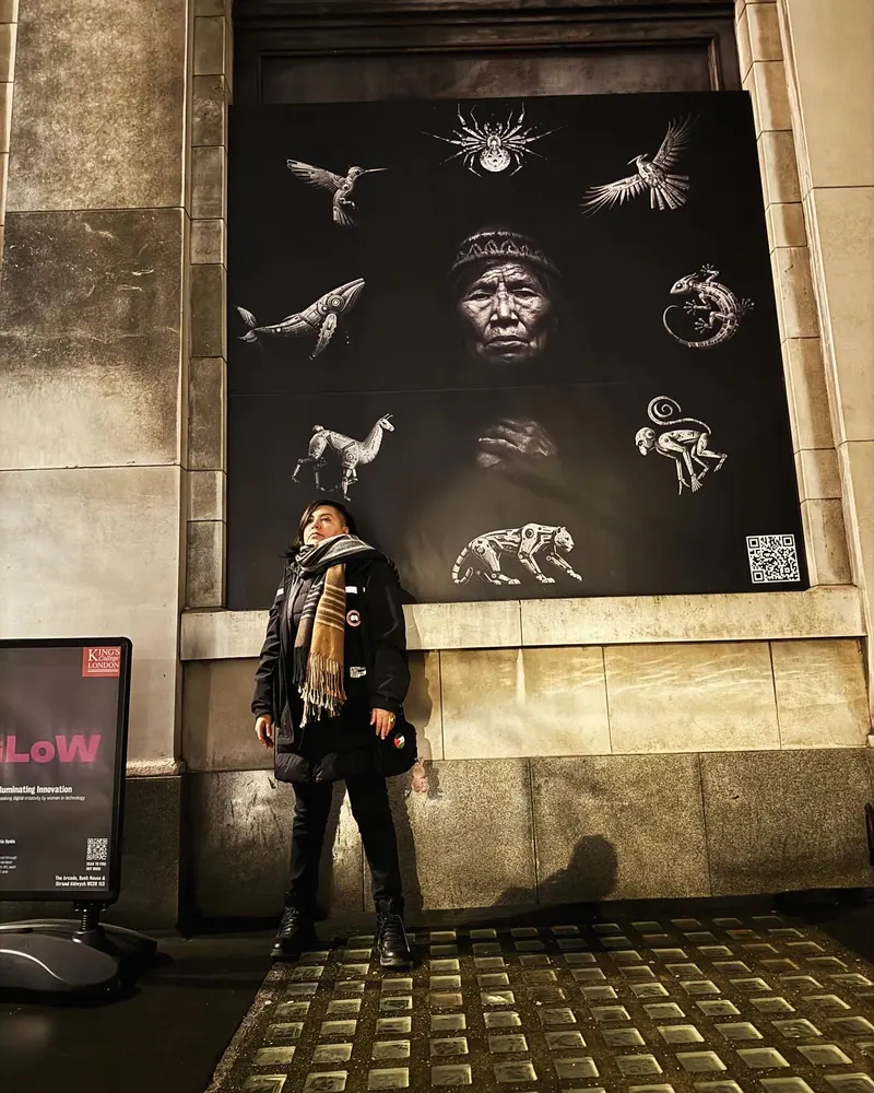 Una persona de pie bajo un gran panel de Las Awichas en un edificio de piedra, de noche. Ocho animales robóticos plateados rodean el rostro de una abuela. El Strand, King's College London, 2024. Personajes animados en 3D de Brian Condori Marza.
