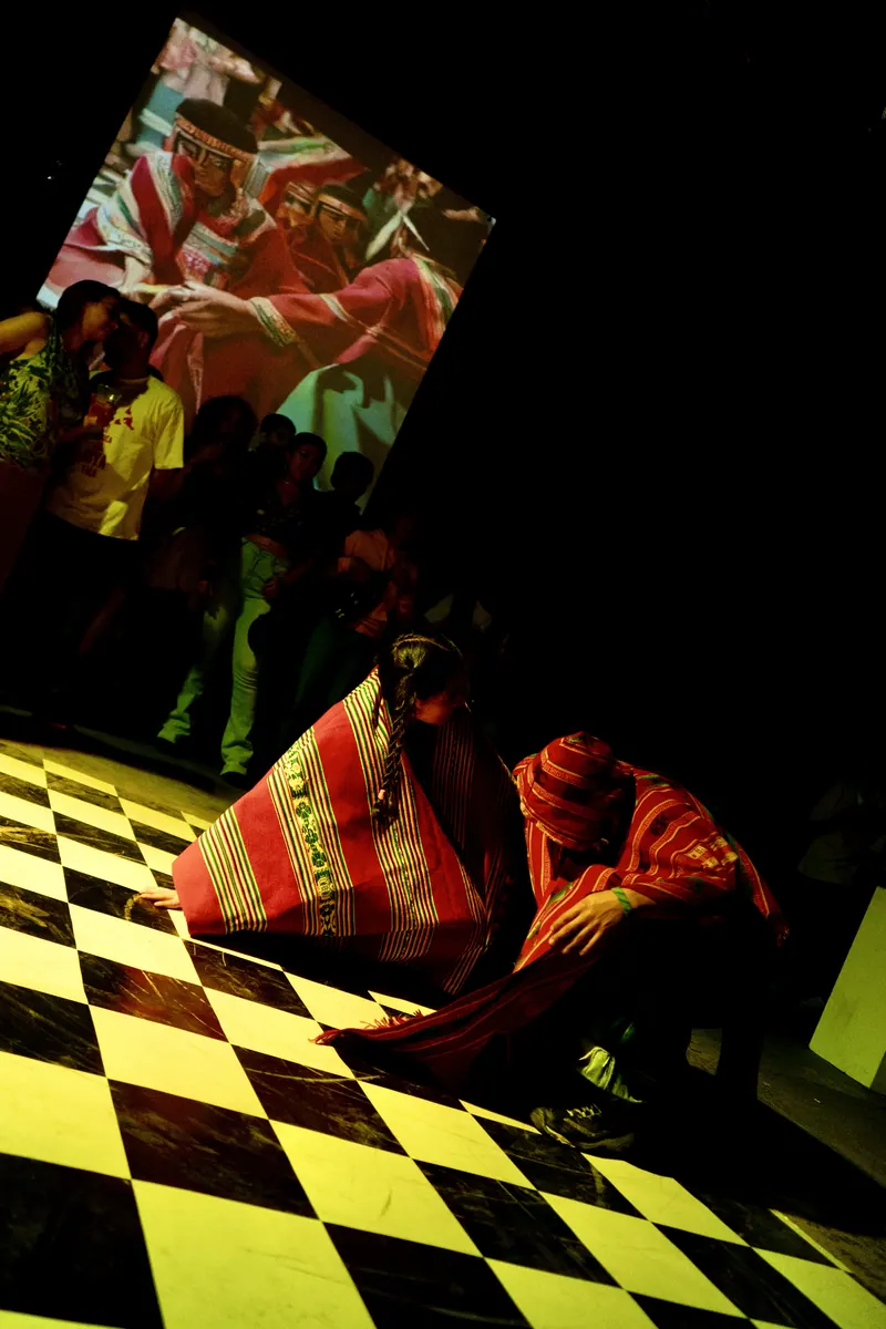 Una figura envuelta en tela roja a rayas sentada sobre un piso a cuadros amarillos y negros. El Martadero, Cochabamba, 2025.