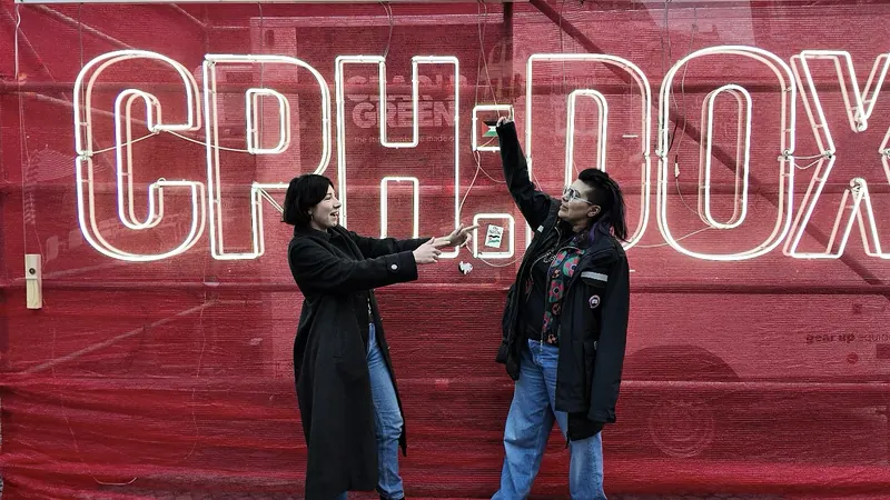 Dos personas bajo un letrero de neón de CPH:DOX en una pared roja. Una levanta un brazo. CPH:DOX, Copenhague.