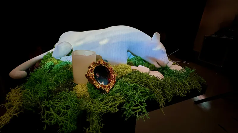 Una escultura blanca de un animal recostada sobre musgo, junto a una vela y un espejo de marco dorado.