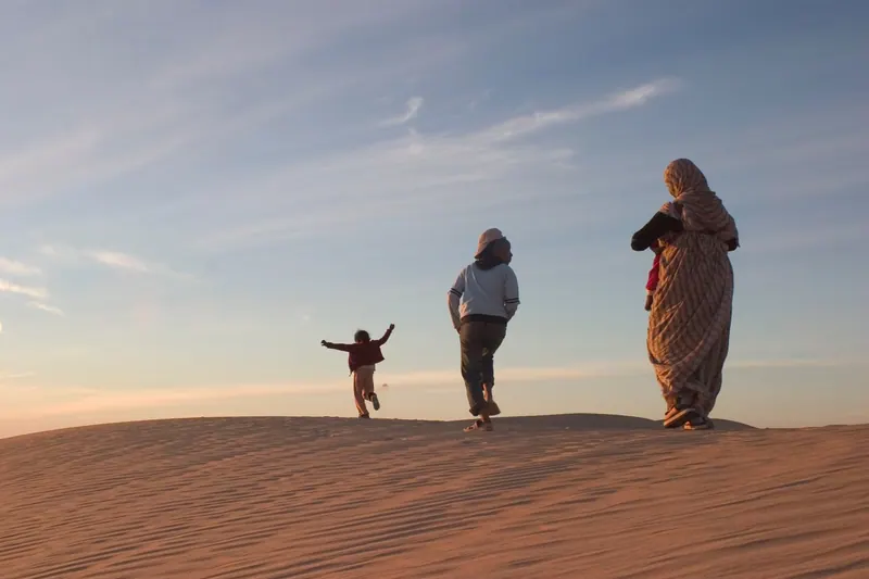 Tres figuras cruzan una duna al atardecer. La más pequeña salta.