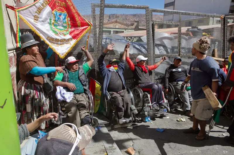 Manifestantes en sillas de ruedas levantan los brazos junto a una malla de alambre, bajo un estandarte bordado.