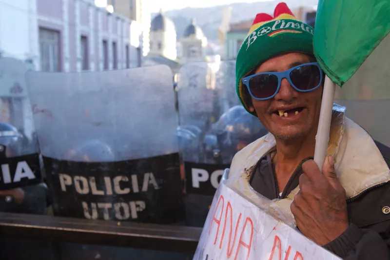 Un hombre con gorro verde y lentes de sol azules frente a escudos policiales.