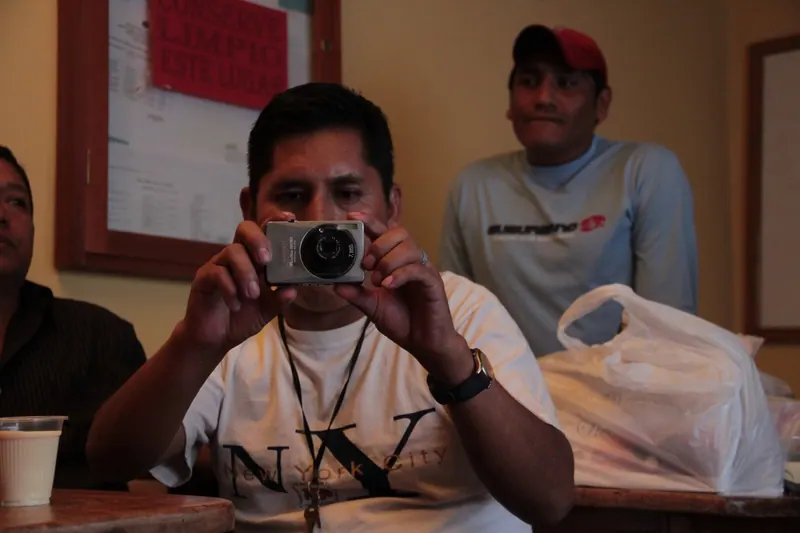 Un hombre apunta una cámara pequeña hacia quien mira. Otro hombre observa desde atrás. Cárcel de San Sebastián, Cochabamba.