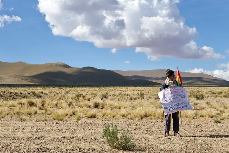 Una persona sola sostiene un cartel escrito a mano y una bandera boliviana en el altiplano. La marcha a La Paz, Bolivia, 2016.