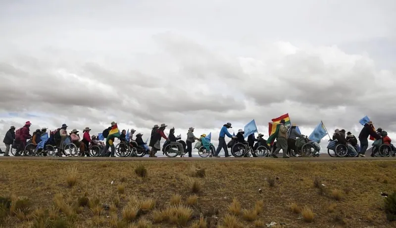 Una larga fila de personas en sillas de ruedas y a pie lleva banderas bolivianas por el altiplano. La marcha a La Paz, Bolivia, 2016.