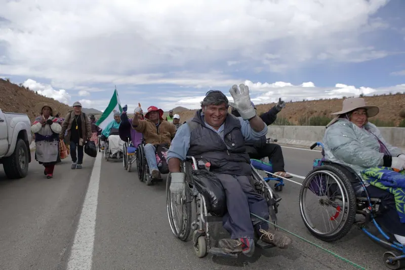 Personas en sillas de ruedas avanzan por un camino de montaña. Una saluda. La marcha a La Paz, Bolivia, 2016.