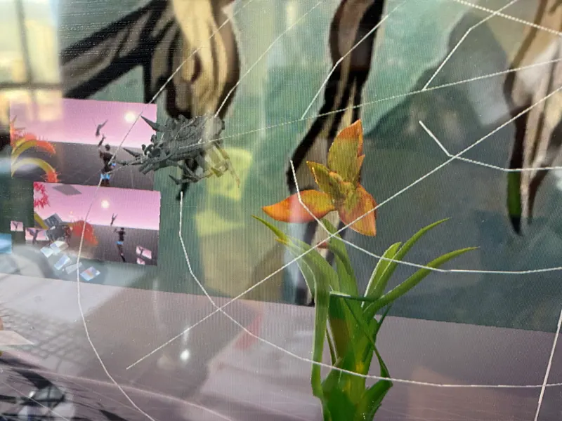 Una escena 3D vista a través de una telaraña: una orquídea naranja, una araña plateada y pantallas flotantes. De la nota de laboratorio La araña aprende a tejer, la gecko aprende a sentir.