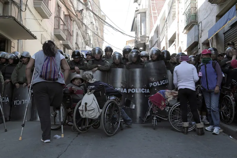 Personas en sillas de ruedas y con muletas frente a una fila de policías antimotines con escudos.