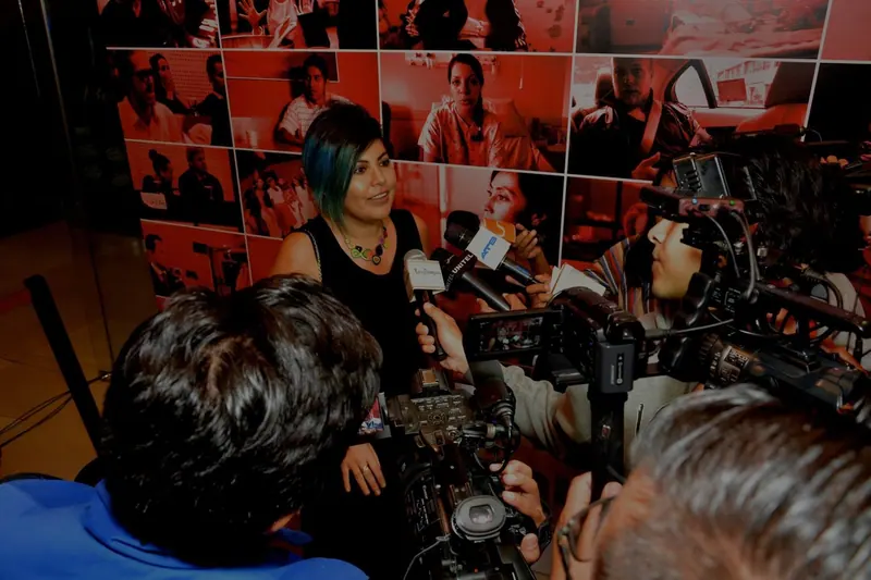 Una mujer habla ante micrófonos y cámaras, frente a una pared de fotografías teñidas de rojo.