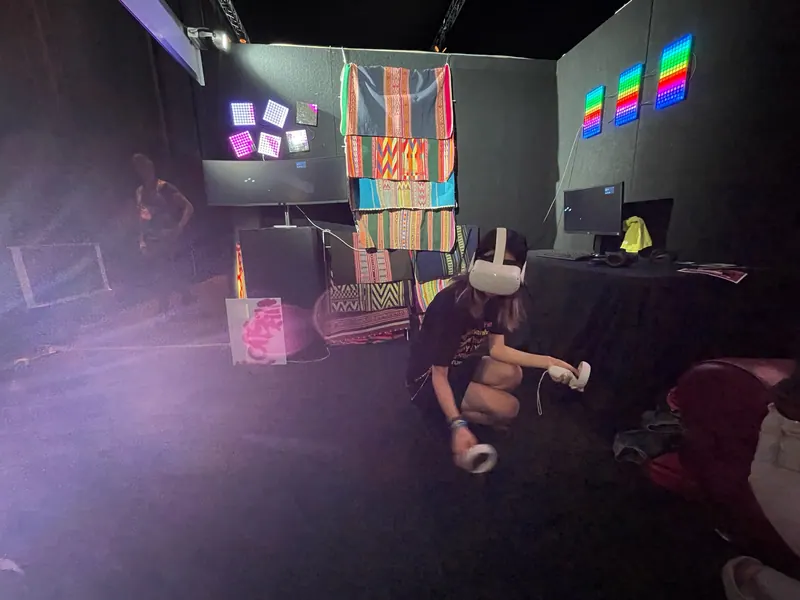 Una persona arrodillada con un visor de VR dentro del stand de Prison X, en luz morada.