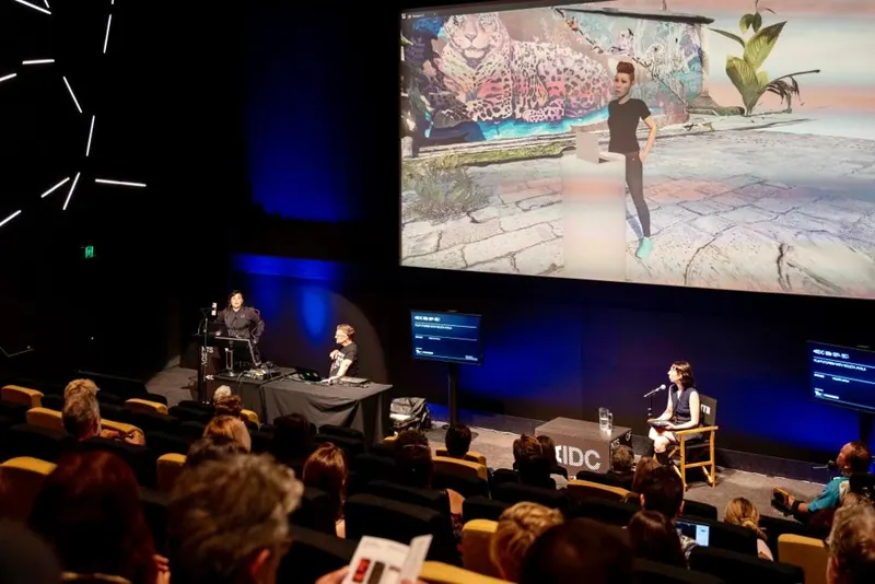 Una charla en un escenario, bajo una pantalla grande que muestra una escena 3D.