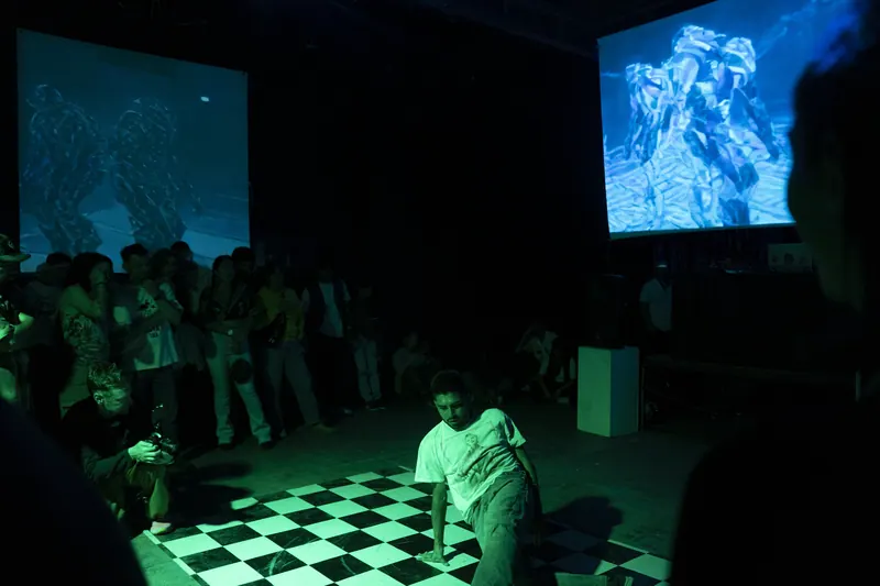 Un bailarín agachado sobre un piso a cuadros, en luz verde. El público lo rodea de pie y dos pantallas muestran criaturas azules. El Martadero, Cochabamba, 2025.