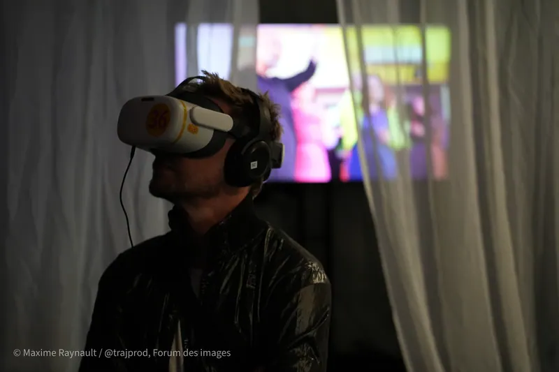 Un hombre con un visor de VR y audífonos frente a una cortina y una pantalla brillante. Forum des images, París.