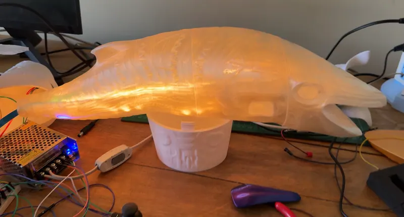 Una ballena translúcida impresa en 3D brilla en naranja sobre una mesa de trabajo. Yakumama en construcción, 2026.
