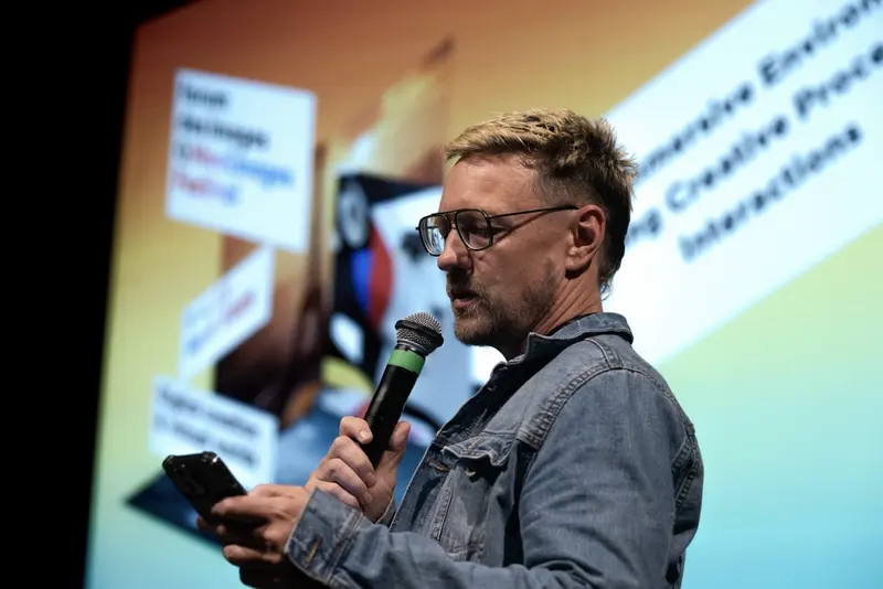 A speaker holds a microphone and a phone on a stage, in front of a projected slide.