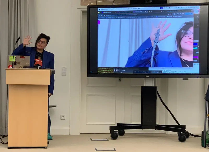 A speaker at a lectern raises a hand beside a large screen. On the screen, a silver gecko sits on that hand.