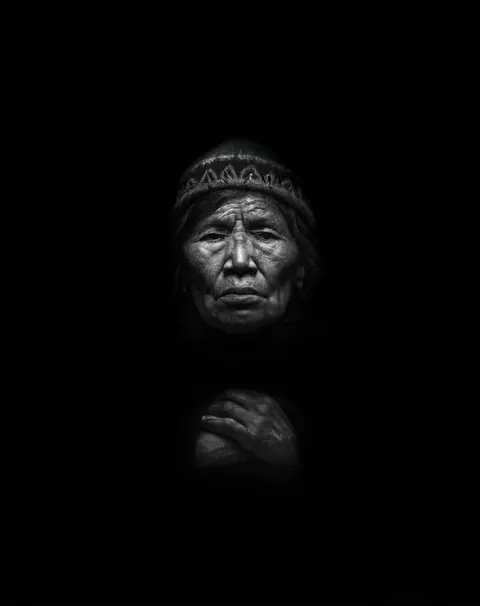 Un retrato de Las Awichas en blanco y negro: una mujer mayor con un gorro tejido y las manos juntas.