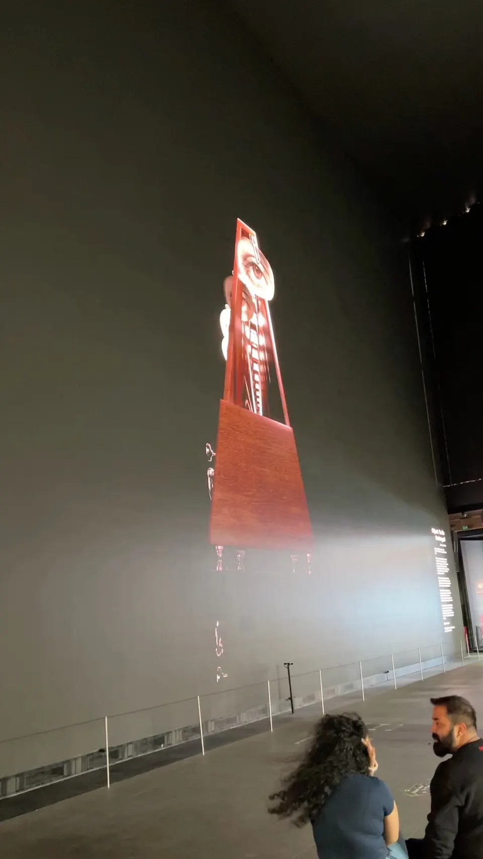 Dos personas miran una forma roja y alta iluminada entre la bruma, en una sala oscura y muy grande.
