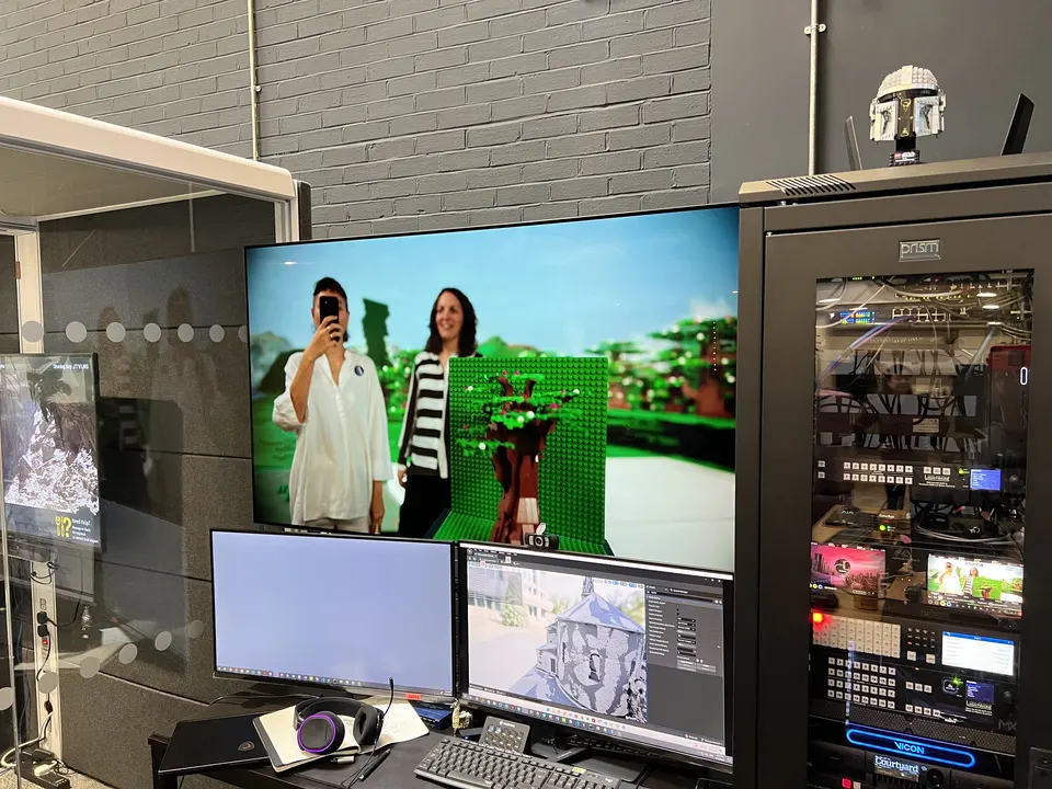 La sala de control de un estudio: una pantalla grande muestra a dos personas delante de una pantalla verde, junto a monitores y un rack de equipos.