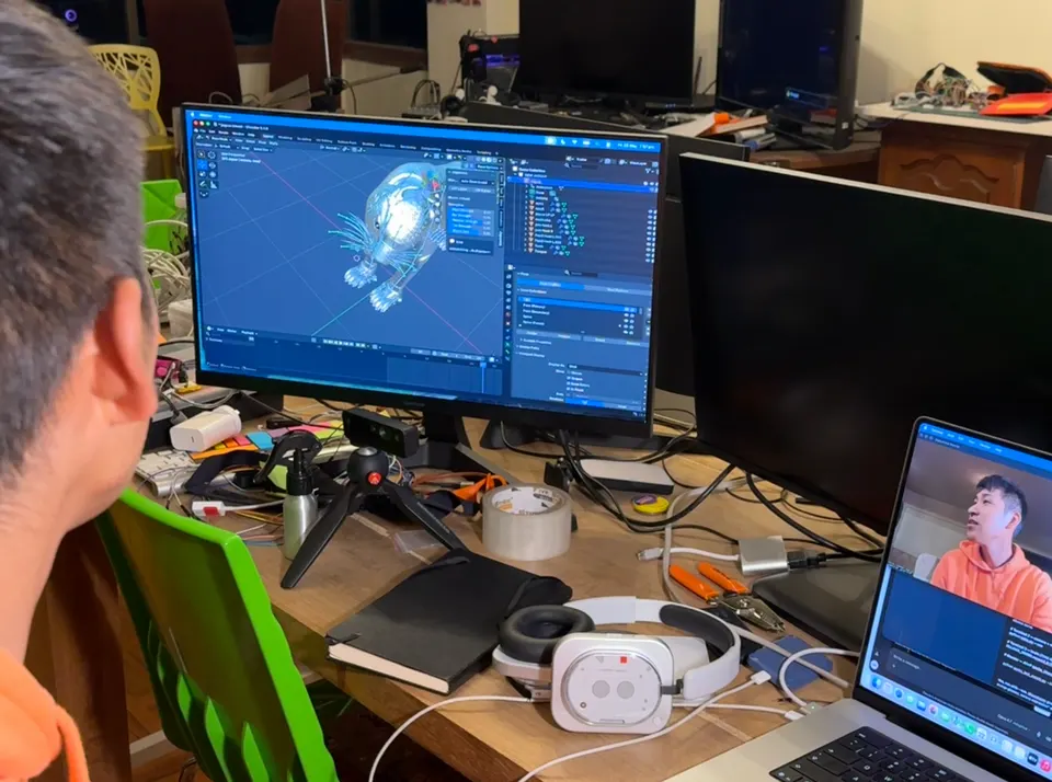 Una persona está sentada ante un monitor que muestra una criatura 3D en Blender. Una laptop muestra una videollamada. Hay un visor de realidad virtual sobre el escritorio.