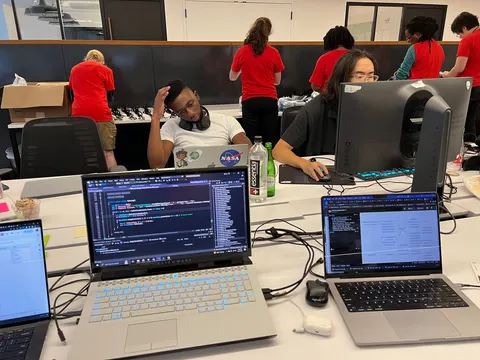 Dos laptops con código sobre un escritorio blanco. Atrás, personas que trabajan y otras de polera roja en un mostrador.