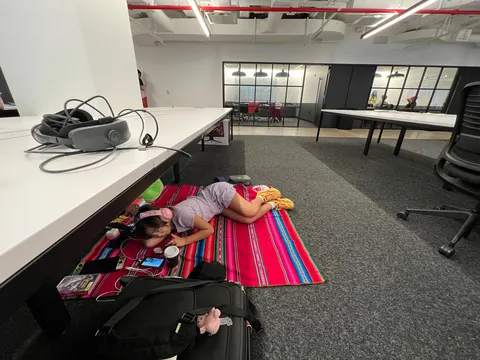 Una niña está echada sobre un aguayo en el piso de la oficina, junto a un escritorio con un visor de realidad virtual.