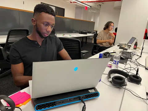 Una persona trabaja en una laptop en un escritorio largo. Otra persona trabaja al fondo.