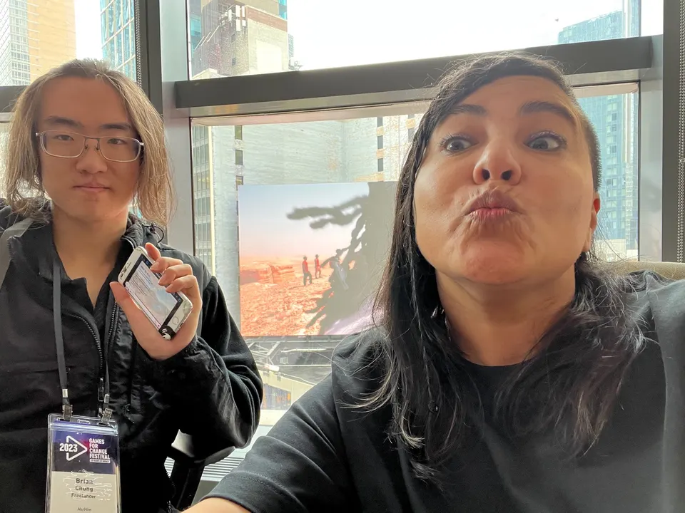 Dos personas se toman una selfie junto a una ventana con torres de la ciudad atrás. Una lleva una credencial del festival y sostiene un celular.
