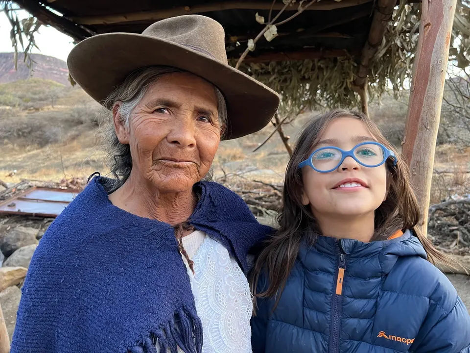 Una mujer mayor con sombrero de fieltro y manta azul está junto a una niña de lentes azules, bajo un techo de ramas.