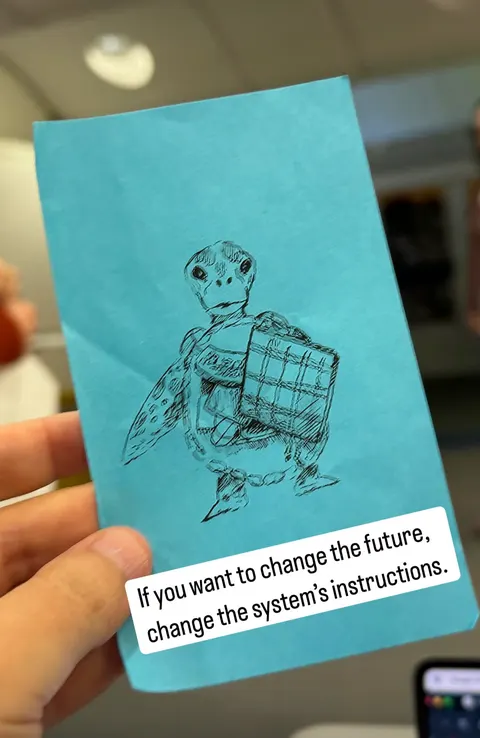 Una mano sostiene una tarjeta azul con el dibujo de una tortuga que carga un bulto. Un texto dice: If you want to change the future, change the system's instructions.