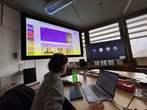 Una persona trabaja en una laptop en una sala de reuniones, frente a una pantalla grande y un monitor con una videollamada.