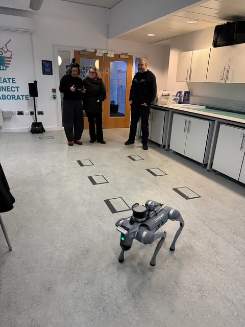 Un robot de cuatro patas está parado en el piso de un laboratorio. Tres personas miran desde el fondo de la sala.