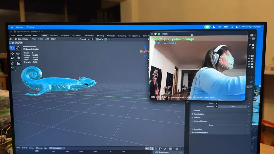 Un monitor muestra un modelo de gecko turquesa en Blender. Una ventana pequeña muestra a una persona con audífonos y puntos de seguimiento en la cara.