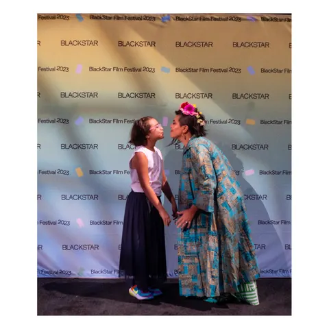 Una niña y una mujer con flores en el pelo se acercan para darse un beso, delante del fondo del BlackStar Film Festival 2023.