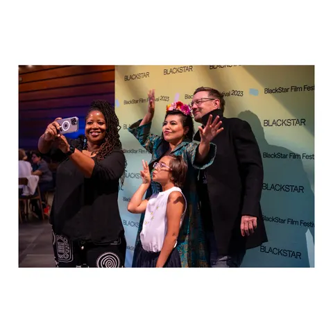 Tres adultos y una niña saludan a un celular para una selfie, delante del fondo de BlackStar.