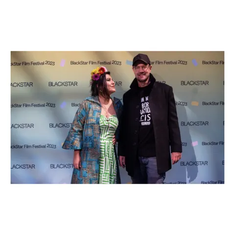Una mujer con flores en el pelo y un hombre de gorra negra posan delante del fondo del BlackStar Film Festival 2023.