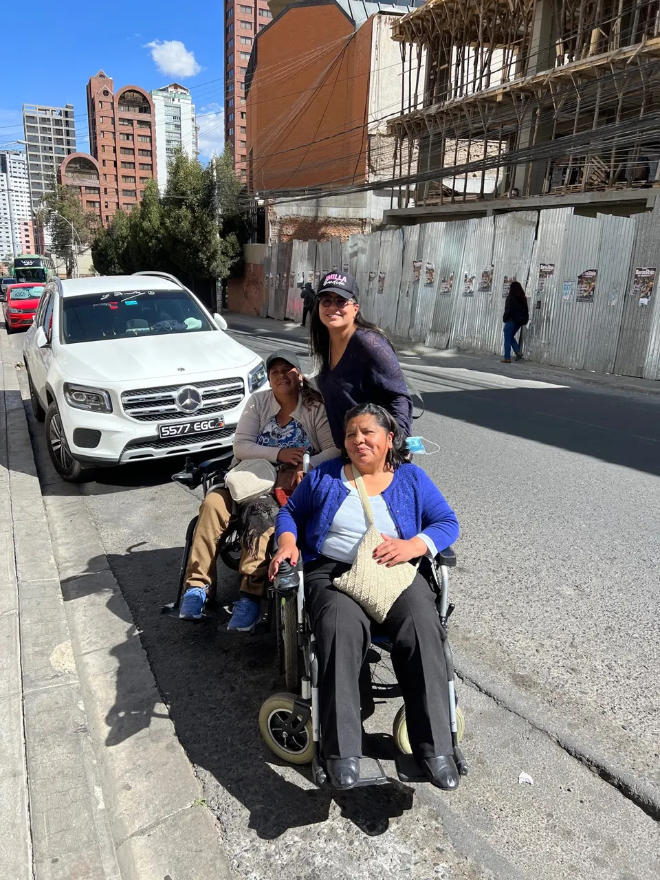 Tres mujeres en una calle empinada junto a un auto estacionado. Dos de ellas usan silla de ruedas.