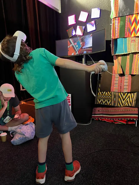 Un niño con un visor de realidad virtual se inclina con un control en cada mano, delante de aguayos y pantallas.