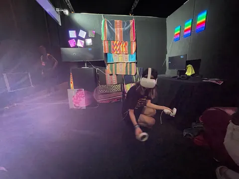 Un espacio de exposición oscuro con aguayos y paneles de luces. Una persona con un visor de realidad virtual está agachada en el piso.