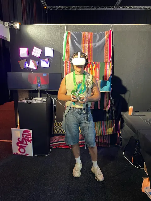 Una persona joven con un visor de realidad virtual sostiene dos controles, delante de aguayos y paneles de luces.
