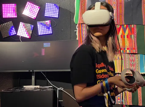 Una persona joven con un visor de realidad virtual sostiene un control. Atrás, paneles de luces rosadas y moradas y aguayos.