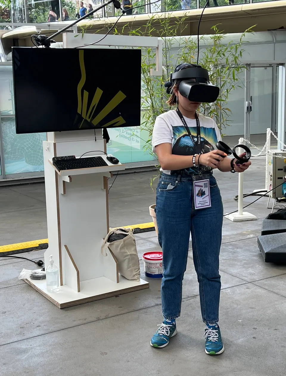 Una persona con un visor de realidad virtual sostiene dos controles junto a una pantalla en un mueble blanco, en un pabellón con paredes de vidrio.